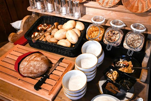 A richly set table with various types of bread and fresh rolls. There are also bowls with muesli, dried fruits, and nuts ready.