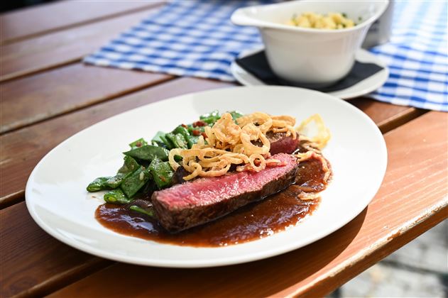 A plate with juicy steak, garnished with crispy onions and fresh vegetables. In the background, there is a bowl with side dishes.
