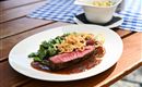 A plate with juicy steak, garnished with crispy onions and fresh vegetables. In the background, there is a bowl with side dishes.
