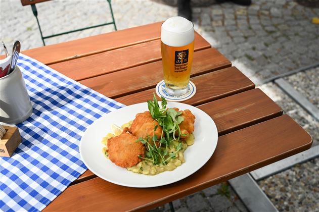 A plate with breaded fish on a creamy side dish, garnished with fresh herbs. Alongside, a glass of beer on a wooden table with a checkered tablecloth.
