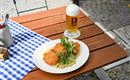 A plate with breaded fish on a creamy side dish, garnished with fresh herbs. Alongside, a glass of beer on a wooden table with a checkered tablecloth.