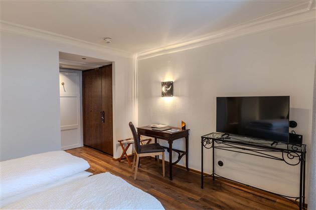 A modern room with a bed, a desk, and a television. The walls are light and the floor is made of wood.