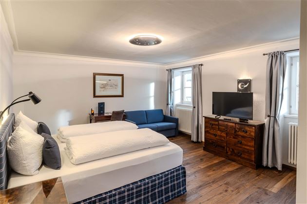 A modern bedroom with two single beds and a cozy sofa. The room is bright and friendly with wooden flooring and large windows.