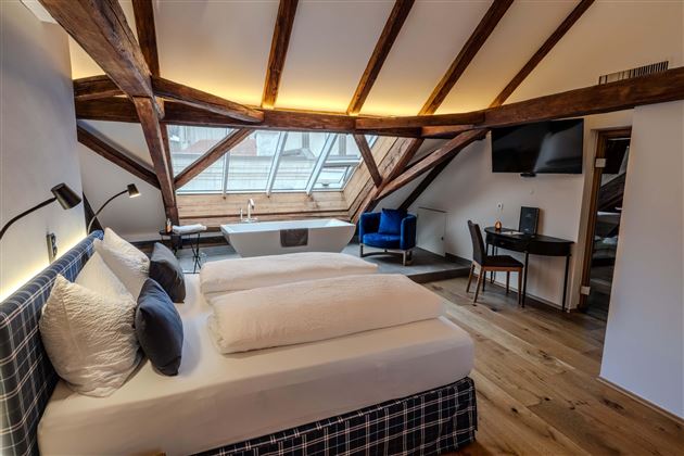 A modern room with wooden beams and large windows. It is equipped with a comfortable bed and a workspace.