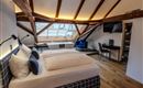 A modern room with wooden beams and large windows. It is equipped with a comfortable bed and a workspace.