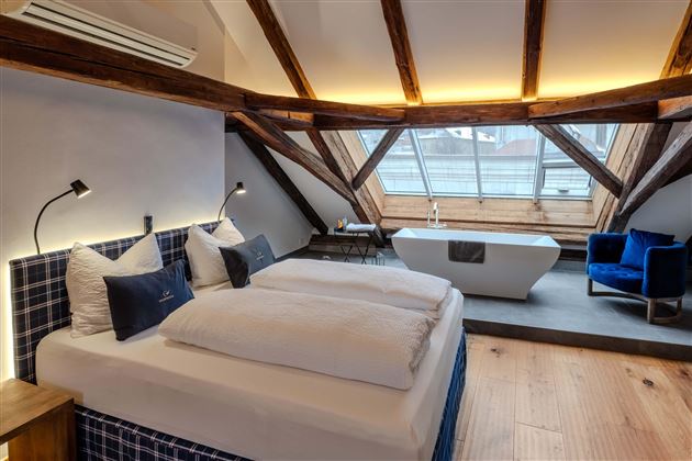 A modern bedroom with a double bed and exposed wooden beams. The large window front provides plenty of natural light and offers a comfortable atmosphere.