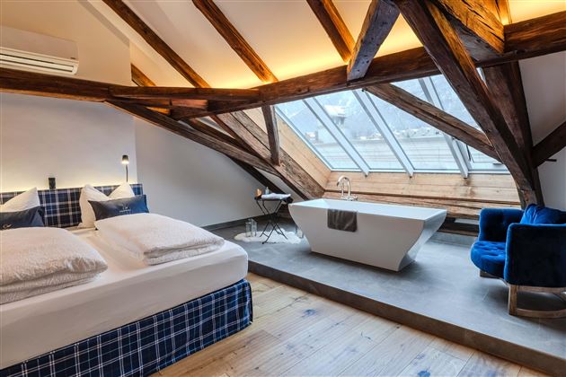 A modern attic room with wooden beams and large windows. There is a comfortable bed, a bathtub, and a cozy armchair.