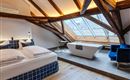 A modern attic room with wooden beams and large windows. There is a comfortable bed, a bathtub, and a cozy armchair.