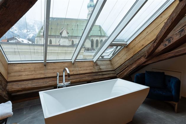 A modern bathroom with a freestanding bathtub and skylights. In the background, a church and the mountains can be seen.