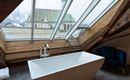 A modern bathroom with a freestanding bathtub and skylights. In the background, a church and the mountains can be seen.