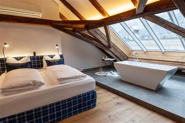 A modern bedroom with a large bed and two pillows. Next to it is a freestanding bathtub and large windows that flood the room with light.