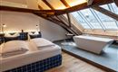 A modern bedroom with a large bed and two pillows. Next to it is a freestanding bathtub and large windows that flood the room with light.