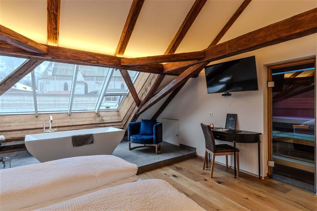 A cozy room with a wooden ceiling and modern furniture. There is a large window, a bathtub, and a television.