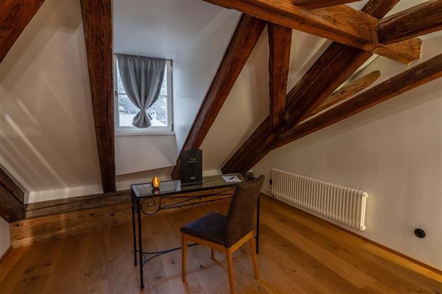 A cozy attic room with visible wooden beams. There is a desk with a chair at the window.