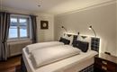 A cozy bedroom with two beds and modern reading lamps. The window lets in plenty of daylight and offers a view outside.