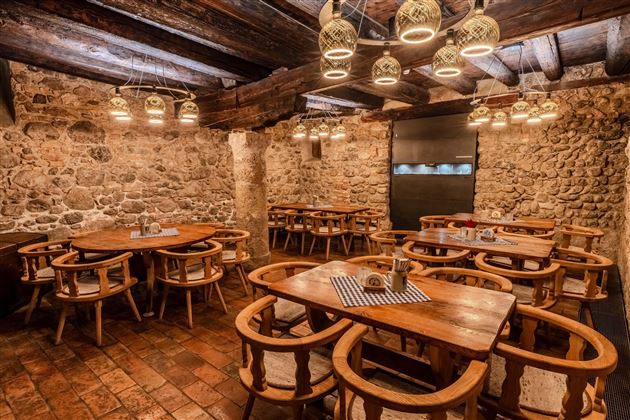 A cozy restaurant with wooden tables and chairs. The walls are made of stones and decorative lamps hang from the ceiling.