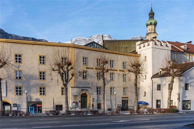 A historic building with a striking clock tower. In the background, the mountains can be seen and the street is lined with trees.