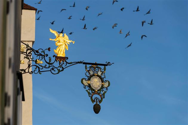 A golden angel on a historical shield under a clear blue sky. Above the shield, many birds are flying.