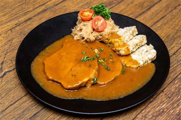 A plate with tender meat in a brownish sauce, accompanied by cabbage and dumplings. Fresh tomato slices and herbs garnish the dish.