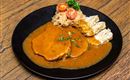 A plate with tender meat in a brownish sauce, accompanied by cabbage and dumplings. Fresh tomato slices and herbs garnish the dish.