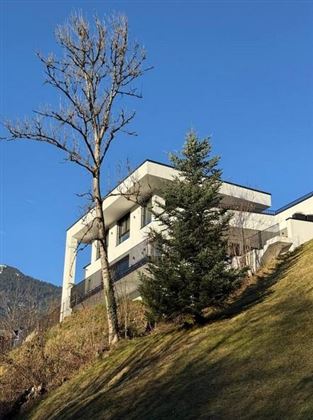 Ein modernes Haus auf einem Hang mit einer schönen Aussicht. Ein kahler Baum und ein immergrüner Baum umgeben das Gebäude.