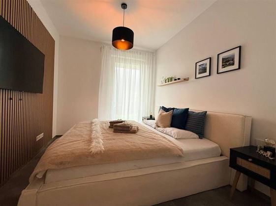 Ein modernes Schlafzimmer mit einem großen Bett und gemütlicher Bettwäsche. Weißes Licht strömt durch die Fenster, während einige Bilder die Wände schmücken.