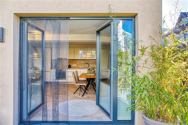 Une porte de terrasse moderne s'ouvre sur une salle à manger lumineuse. Au premier plan, des plantes de bambou sont visibles, créant une atmosphère naturelle.