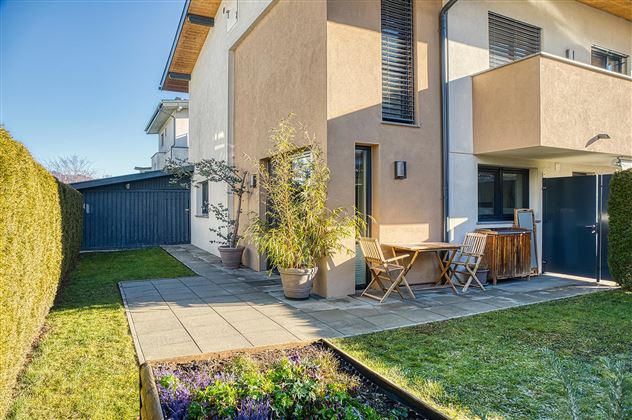 Une maison moderne avec une terrasse cozy et un petit jardin.  
Des parterres de fleurs et une haie verte entourent l'extérieur.