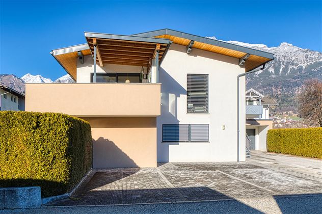 Une maison moderne avec une véranda en bois et de grandes fenêtres. À l'arrière-plan, on peut voir des montagnes enneigées.