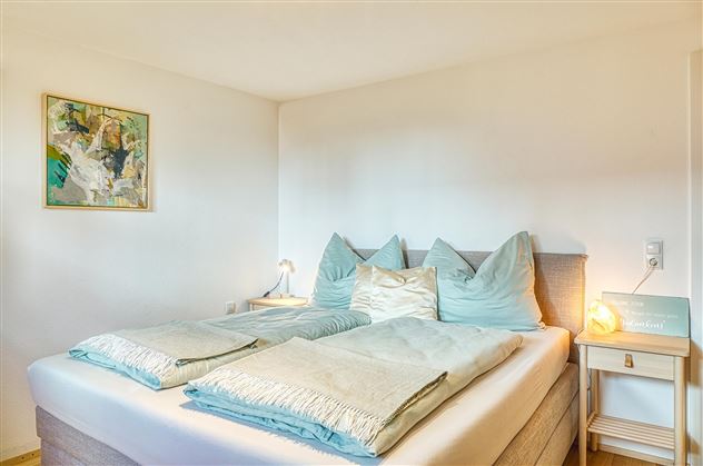 Une chambre confortable avec un grand lit et de jolis coussins verts. Les murs sont clairs et il y a une image ainsi qu'une lampe de chevet.