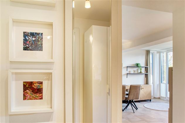 Une entrée moderne avec des images colorées sur le mur et un placard blanc. À l'arrière-plan, un salon confortable avec une grande baie vitrée est visible.