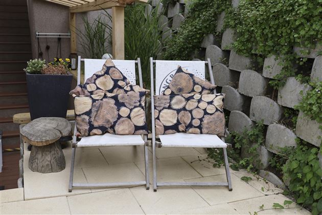 Two lounge chairs with cushions in a wooden design stand next to a small table. In the background, green plants and a tiled wall can be seen.