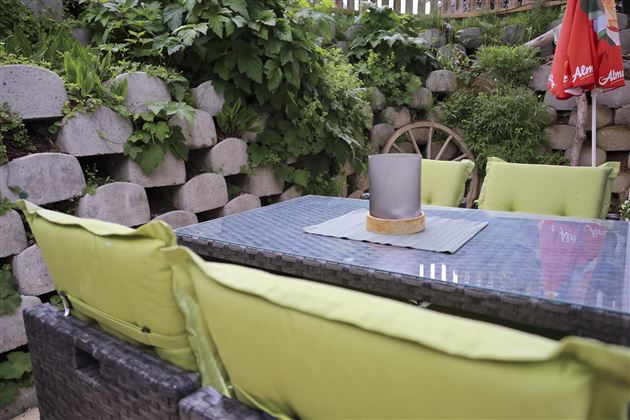 A cozy garden area with a table and green cushions. In the background, plants and a stone wall are visible.