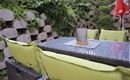 A cozy garden area with a table and green cushions. In the background, plants and a stone wall are visible.