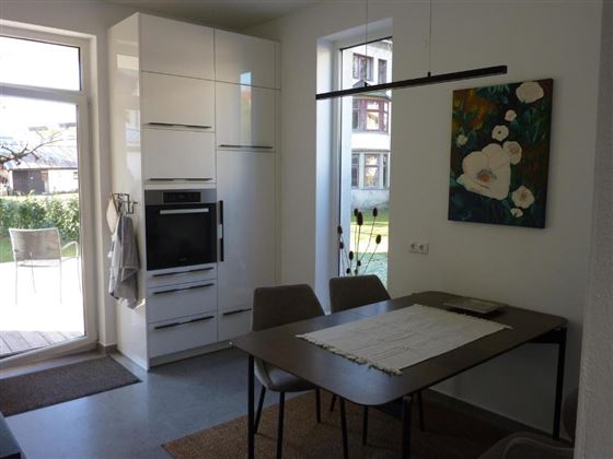 A modern kitchen with white furniture and a dining area. 

A lot of daylight comes through the windows and the view overlooks a beautiful garden.