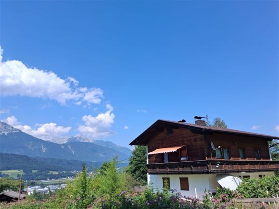 Een traditioneel houten huis met een balkon, omringd door groene natuur. Op de achtergrond zijn indrukwekkende bergen zichtbaar onder een heldere blauwe lucht.