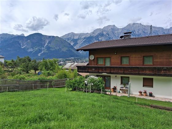 Een gezellig houten huis met balkon en tuin. Op de achtergrond zijn indrukwekkende bergen en een groen landschap te zien.