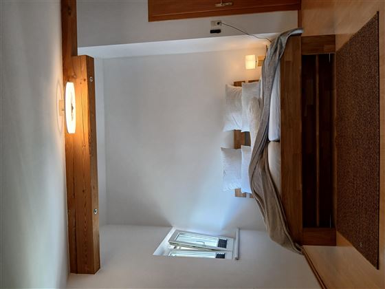 Ein modernes Schlafzimmer mit einem Doppelbett und weißen Kissen. Es gibt holzfarbene Akzente und eine große Fensterfläche, die Tageslicht hereinlässt.