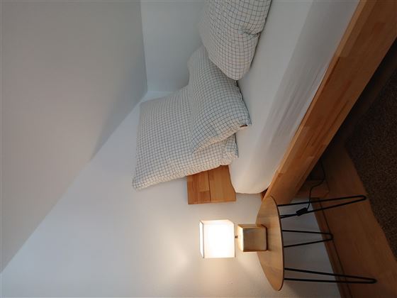Ein einfaches Schlafzimmer mit einem Bett und zwei Kopfkissen. Neben dem Bett steht ein Nachttisch mit einer Lampe.