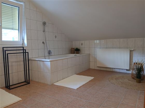Ein geräumiges Badezimmer mit einer Badewanne und hellen Fliesen. Es gibt einen Heizkörper und eine Fenster mit natürlichem Licht.