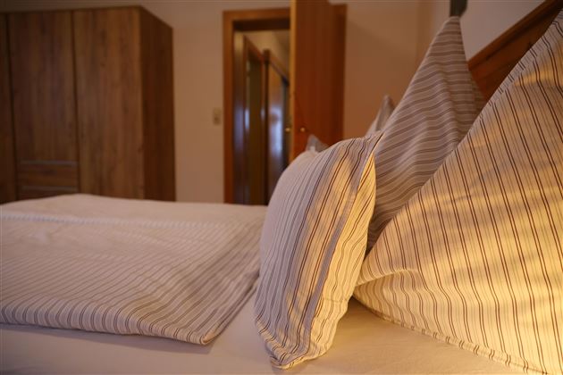 Ein gemütliches Schlafzimmer mit einem Bett und gestreiften Kopfkissen. Im Hintergrund sind warme Holztöne und eine offene Tür zu sehen.