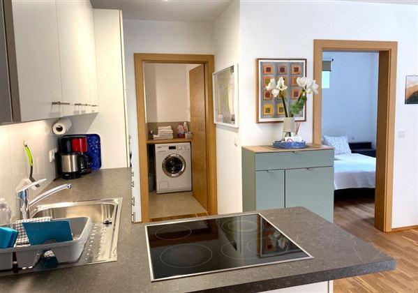 Een moderne keuken met een kookeiland en een achterliggende wasruimte. In de aangrenzende kamer is een gezellige slaapkamer zichtbaar.