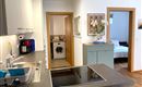 Een moderne keuken met een kookeiland en een achterliggende wasruimte. In de aangrenzende kamer is een gezellige slaapkamer zichtbaar.