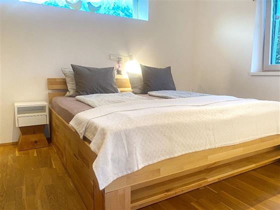 Een gezellige slaapkamer met een groot bed en grijze kussens. De kamer is licht en opgeruimd, met houten vloeren en een raam dat daglicht binnenlaat.