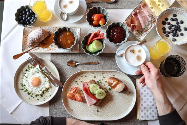 Ein reichhaltiges Frühstück mit verschiedenen Speisen wie Eiern, Brot, Aufschnitt und frischem Obst. Dazu gibt es Kaffee und verschiedene Marmeladen auf einem schön gedeckten Tisch.