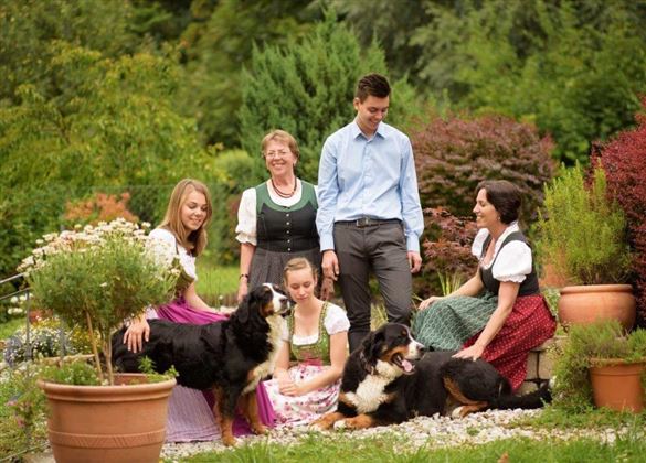 Eine Gruppe von fünf Personen in traditionellen Trachten sitzt im Garten. Zwei Hunde spielen in der Nähe, umgeben von bunten Pflanzen und Blumen.