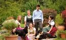 Eine Gruppe von fünf Personen in traditionellen Trachten sitzt im Garten. Zwei Hunde spielen in der Nähe, umgeben von bunten Pflanzen und Blumen.