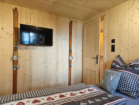 Ein gemütliches Zimmer mit Holzwänden und einem Fernseher an der Wand. Auf dem Bett liegen dekorative Kissen und eine ansprechende Bettdecke.