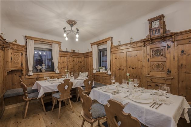 A cozy dining room with wooden paneling and elegantly set tables. Windows with curtains and warm lighting create an inviting atmosphere.