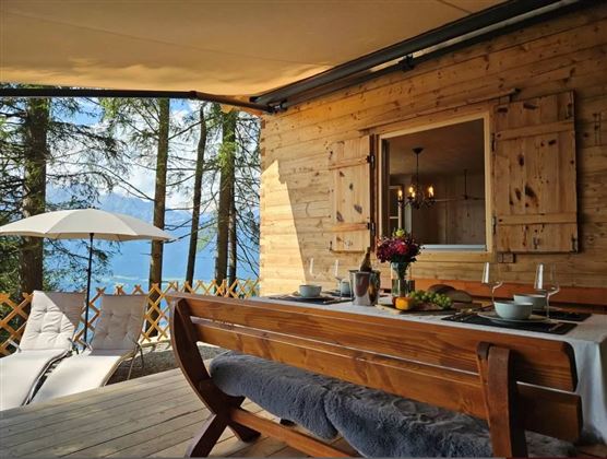 Eine gemütliche Terrasse mit einem Holztisch und zwei Liegen. Im Hintergrund sind Bäume und ein schöner Ausblick auf einen See.